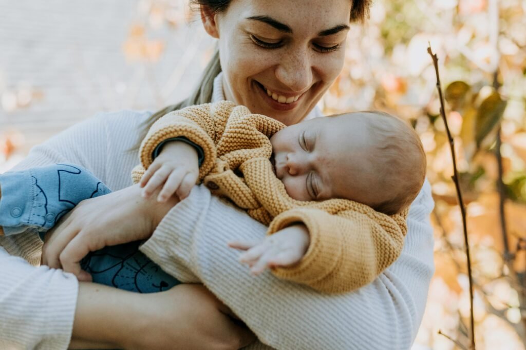 Parent smiling while holding a peacefully sleeping baby outdoors on a warm autumn day, representing calm and connected parenthood in a cozy, natural setting.