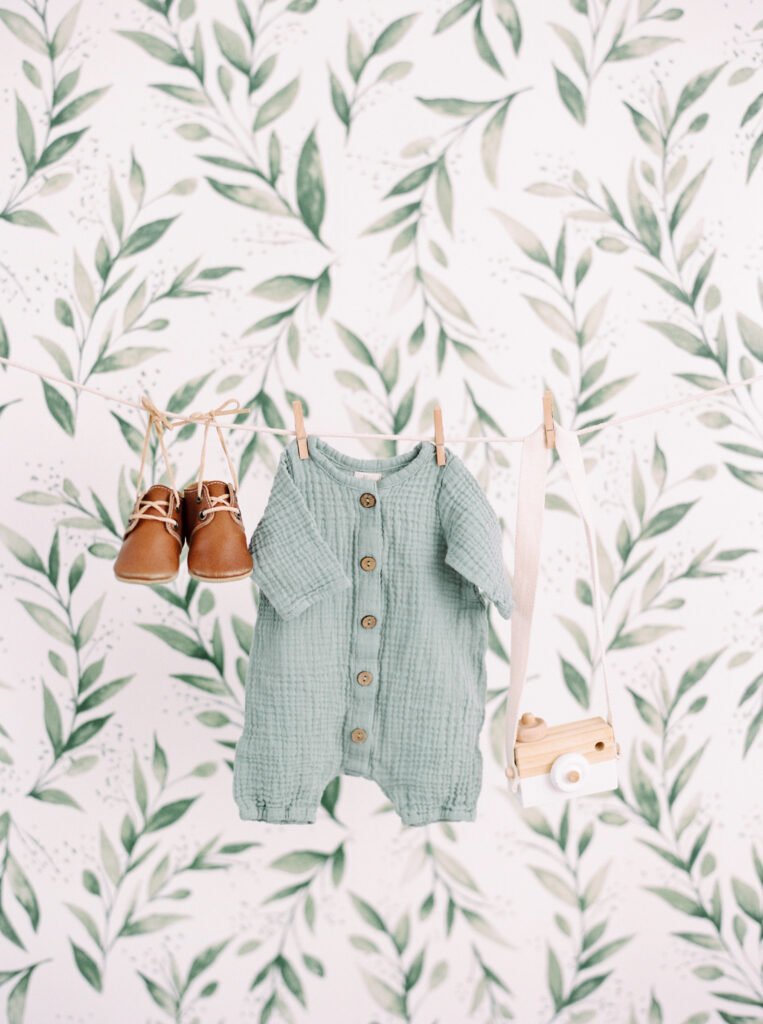 Neutral baby outfit with sage green romper, tan leather shoes, and wooden toy camera hanging on a clothesline in front of leafy wallpaper β LullaCozy calm nursery inspiration.